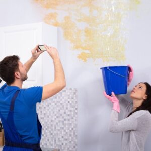 woman holds blue bucket while plumber takes photo of water damage